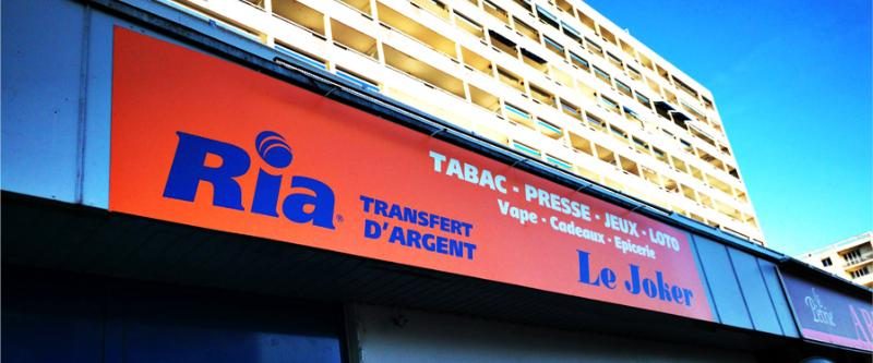 Enseigne lumineuse rouge d'un tabac-presse Ria posée sur une façade d'immeuble
